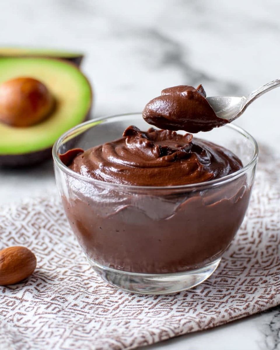 A clear glass bowl with a wide rim sits on a white marbled surface. Inside the bowl, thin swirls and smears of dark brown chocolate cream coat the inner sides, showing texture and light reflections. A silver spoon rests inside, angled with its handle leaning out of the bowl. In the blurred background, there is a small black bowl holding a brown round fruit and a few green leaves. The colors contrast softly with the white marbled surface. photo taken with an iphone --ar 4:5 --v 7