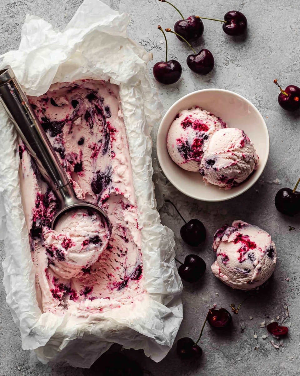 A white rectangular container lined with crumpled parchment paper holds creamy cherry ice cream, pink with deep red cherry swirls spread unevenly across the surface. A metal ice cream scoop rests inside, showing some of the ice cream removed in a smooth round shape. Next to it, a white bowl contains two scoops of the same swirled ice cream, both showing a mix of light pink and dark cherry red colors. Scattered around the scene on a white marbled surface are whole dark cherries with stems. Photo taken with an iphone --ar 4:5 --v 7