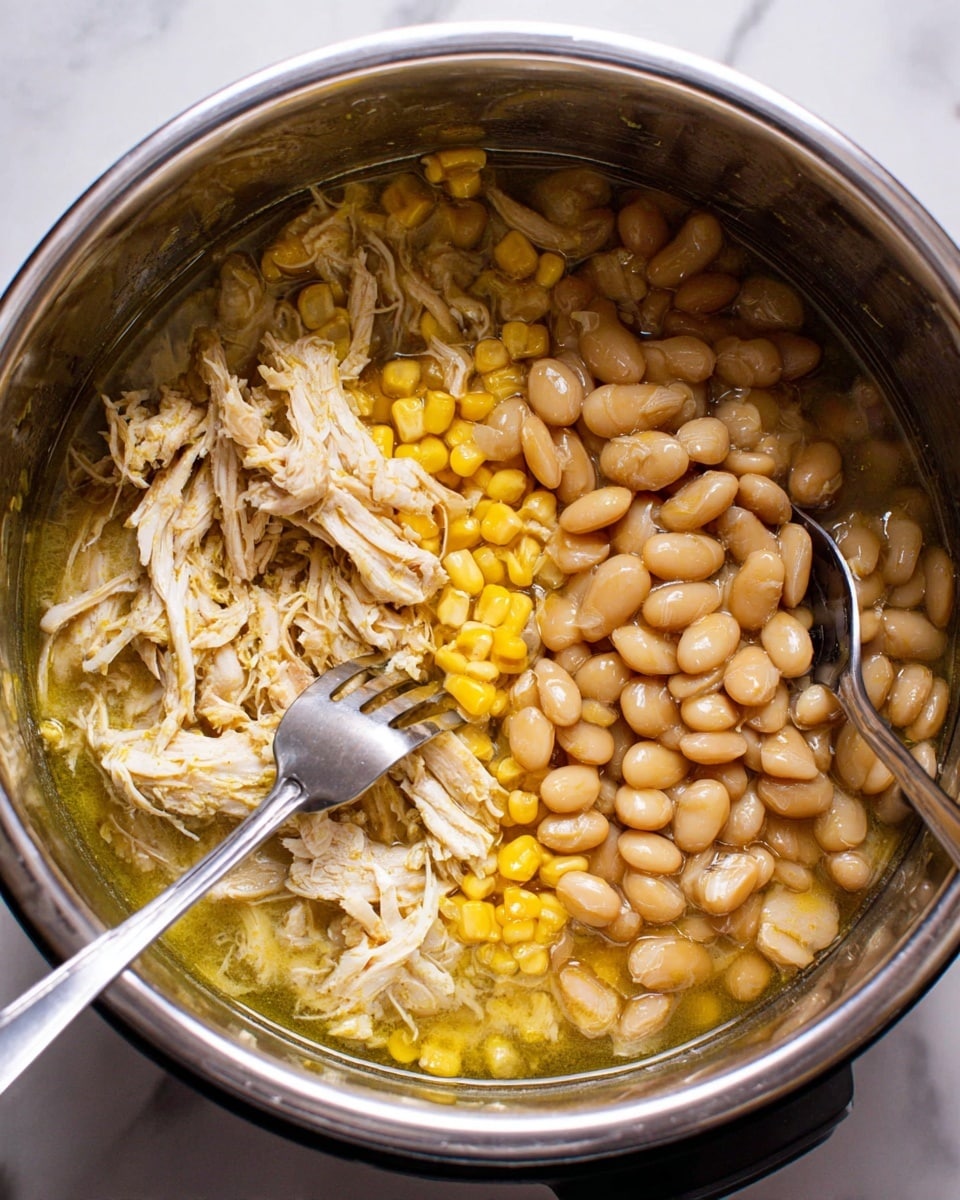 A close-up view inside a shiny silver pot shows a dish with three main layers: on the left and center, there is light yellow shredded chicken mixed with bright yellow corn kernels, all in a light yellow-green broth; on the right side, there are small, light brown beans sitting in the same broth, filling the pot's curved interior. Two silver forks rest diagonally inside the pot, one on the left and one on the right, partly submerged in the food. The pot sits on a white marbled surface. photo taken with an iphone --ar 4:5 --v 7