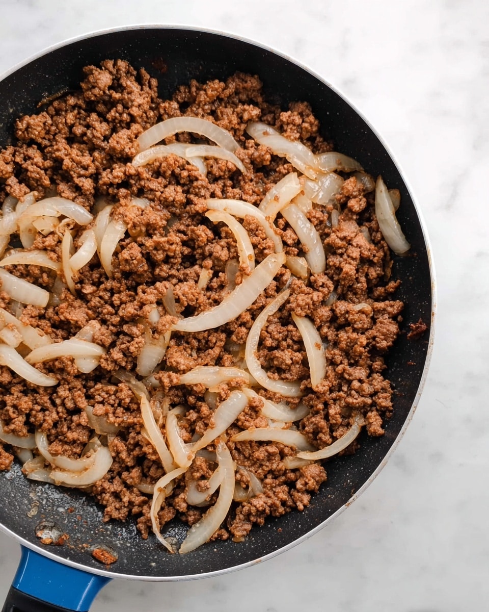 A close-up image of a black frying pan filled with cooked ground meat mixed with translucent, soft cooked onion slices. The meat is brown and crumbly, evenly spread across the pan's surface with the light-colored onions scattered throughout, adding texture contrast. The pan handle is blue and black, resting on a white marbled surface. The scene shows the ingredients well cooked but not browned too much. Photo taken with an iphone --ar 4:5 --v 7