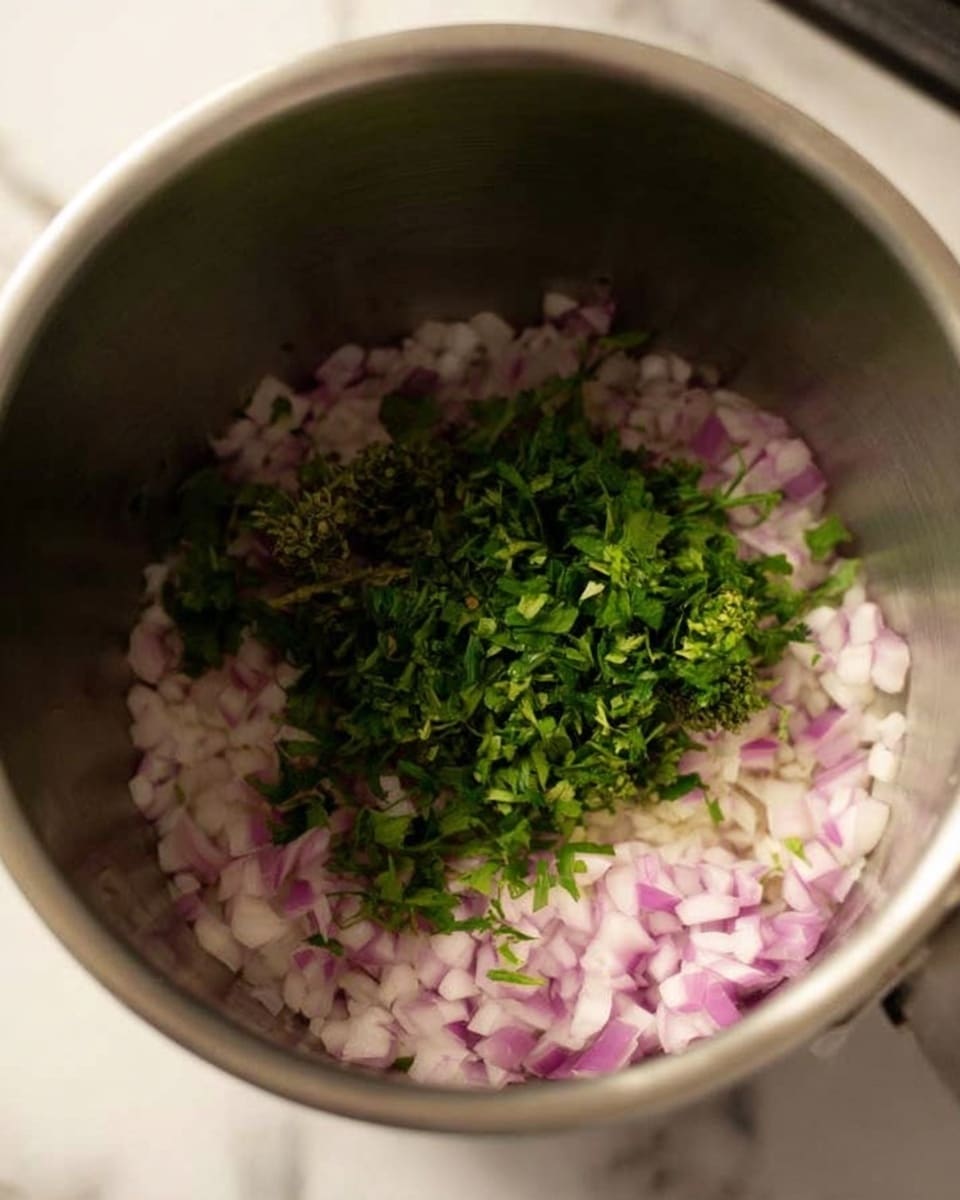 Inside a stainless steel pot, there is a layer of finely chopped light purple onions spread evenly at the bottom, topped with a small bunch of bright green chopped fresh herbs placed in the center. The pot sits on a stove with a white marbled surface barely visible around it. photo taken with an iphone --ar 4:5 --v 7