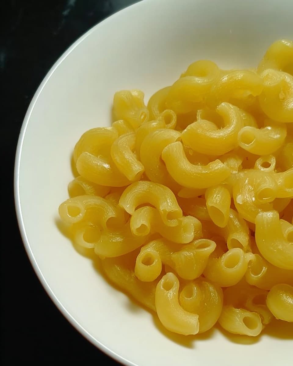 The image shows a close-up of a white bowl filled with cooked macaroni pasta. The macaroni pieces are yellow, smooth, and curved, tightly packed inside the bowl. The bowl is placed on a white marbled surface, and the background is dark, making the yellow pasta stand out clearly. photo taken with an iphone --ar 4:5 --v 7