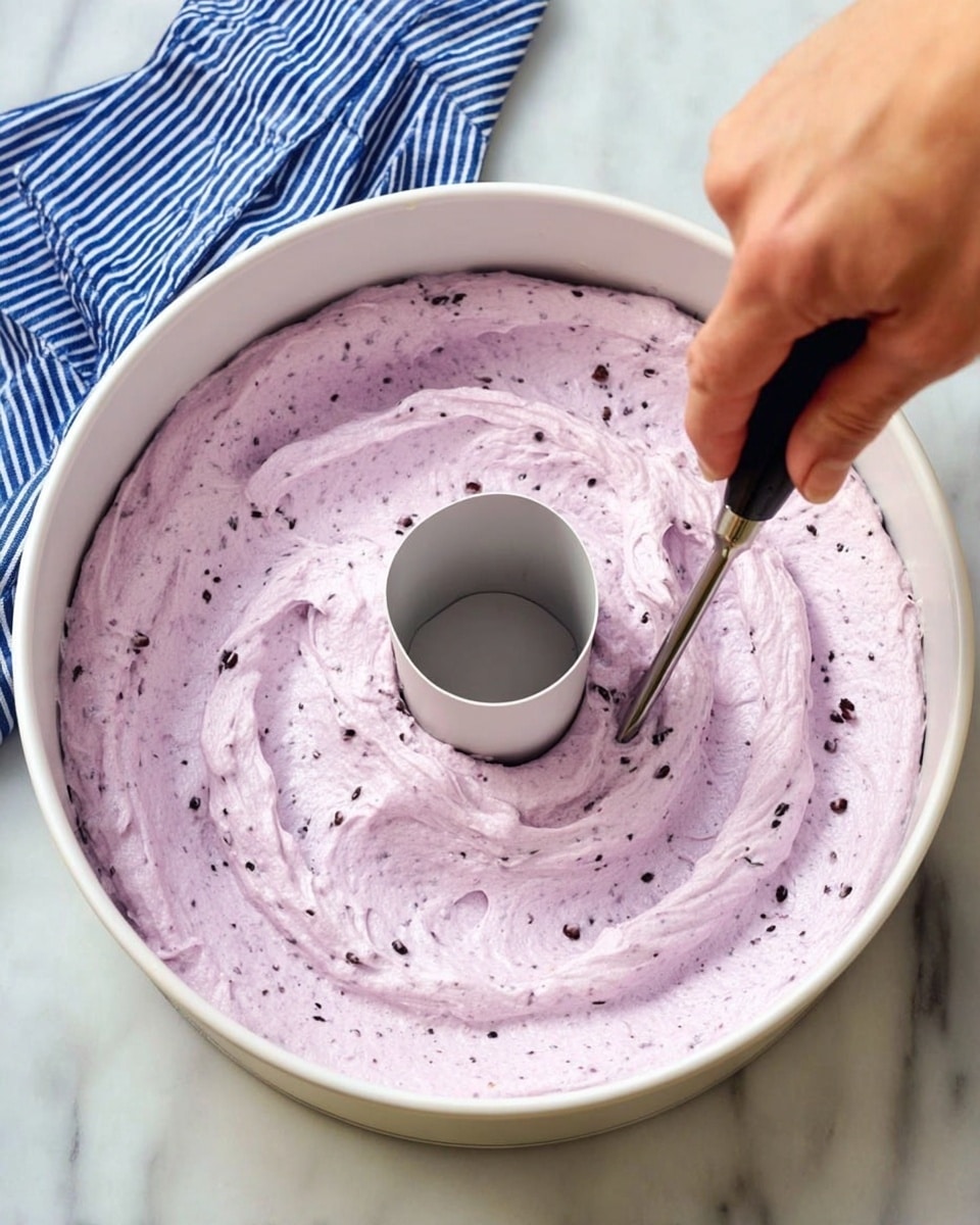 A white tube pan filled with light purple fluffy batter that has small dark bits mixed in, giving it a speckled texture. The batter looks smooth and creamy, swirled evenly around the center tube of the pan. A woman's hand is holding a small black-handled tool, gently poking into the batter near the edge of the pan. The pan is placed on a white marbled surface, and a blue cloth with white stripes is partially visible in the background. Photo taken with an iphone --ar 4:5 --v 7