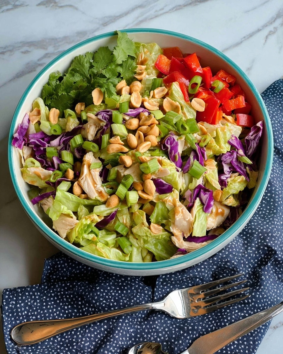 A fresh salad is shown in a white bowl with a thin turquoise rim, placed on a white marbled surface. The salad has many layers, starting with a base of light green lettuce and purple cabbage pieces, mixed with small white chicken pieces scattered all around. On top, there are bright green sliced scallions and chopped light green celery stalks. Bright red bell pepper chunks add a pop of color, and the entire salad is sprinkled with whole golden peanuts. Fresh green cilantro leaves are spread over the salad. A shiny metal serving spoon and fork are on a folded dark blue cloth with small white dots next to the bowl. The overall composition is bright and colorful, emphasizing freshness and texture. Photo taken with an iphone --ar 4:5 --v 7