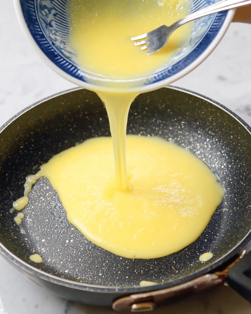 A close-up image shows a light yellow beaten egg mixture being poured from a blue and white patterned bowl into a black non-stick frying pan with small oil drops glistening inside. The yellow egg liquid forms a smooth, even layer spreading over the pan's dark speckled surface. A silver fork is held inside the bowl, guiding the flow. The entire scene is set against a white marbled background. photo taken with an iphone --ar 4:5 --v 7