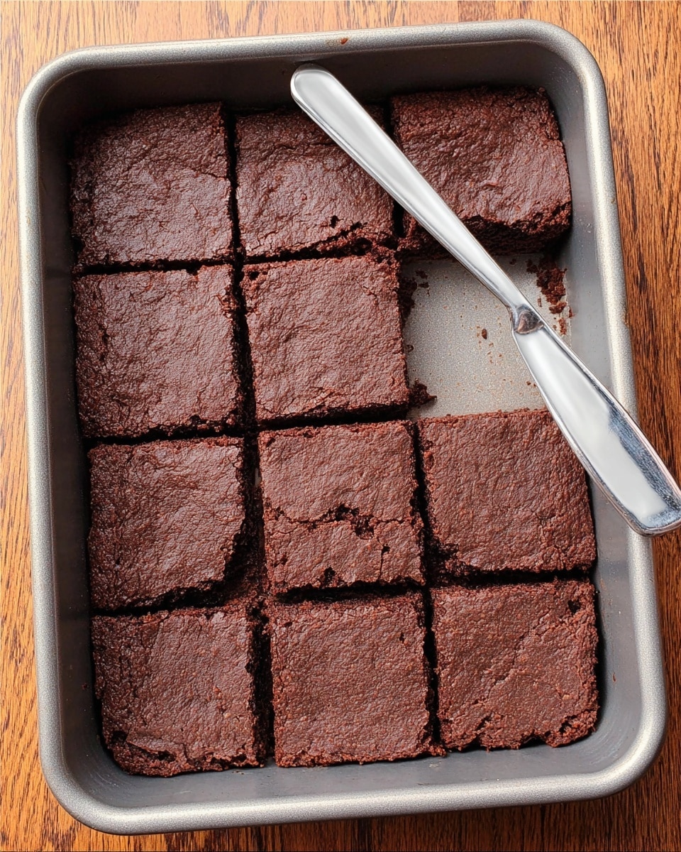 A metal baking pan filled with nine square pieces of dark chocolate brownies, one piece is missing from the bottom left corner, showing the pan’s silver inside. The brownies have a slightly rough texture with small cracks and a matte finish. A metal butter knife with a shiny surface is resting diagonally on the top right corner of the pan. The pan is placed on a wooden surface with visible grain patterns. photo taken with an iphone --ar 4:5 --v 7