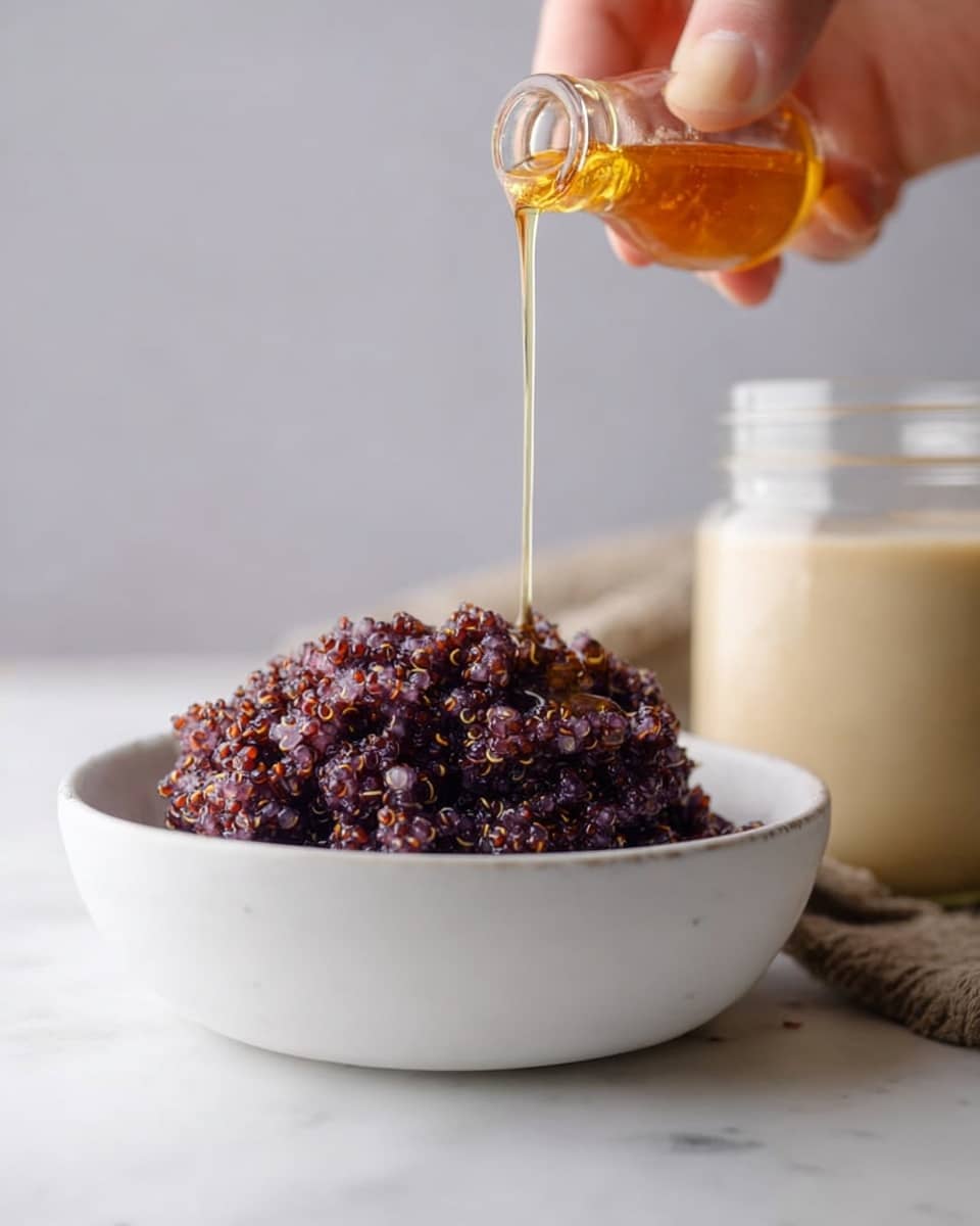 A close-up showing a woman's hand pouring golden honey from a small clear bottle onto a mound of dark purple cooked quinoa inside a simple white bowl. The quinoa layer has a grainy, moist texture with tiny, translucent grains visible. In the soft background, there is a blurred transparent glass jar filled with a creamy light beige liquid. The whole scene sits on a white marbled surface with soft, neutral lighting emphasizing the textures and colors. Photo taken with an iphone --ar 4:5 --v 7