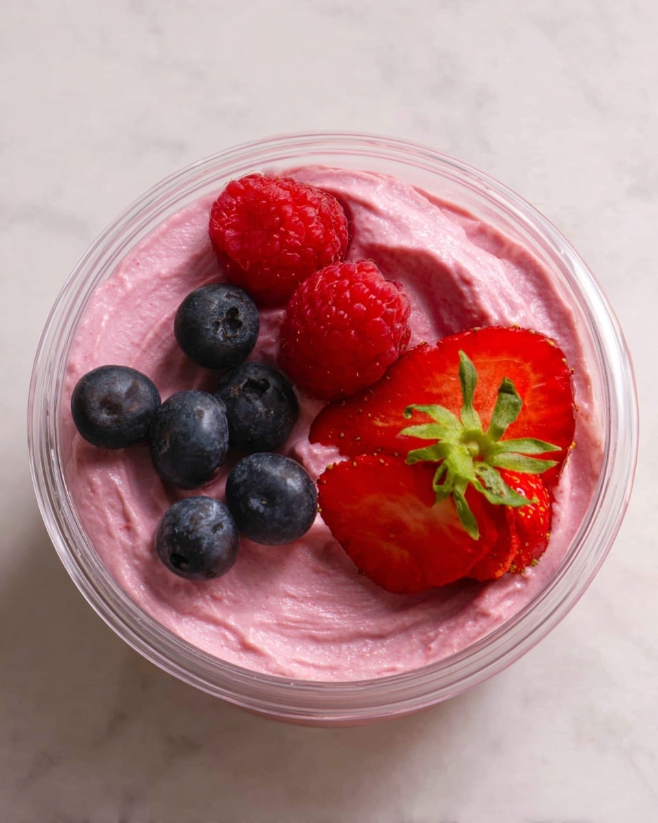 A clear glass bowl sits on a white marbled surface, filled with two scoops of pink berry ice cream, showing a creamy texture with small uneven spots. Mixed berries surround the ice cream inside the bowl – a halved strawberry with visible seeds and green leaves on the side, dark blue blueberries, and bright red raspberries with their bumpy surface. Outside the bowl, in front, there are more scattered whole raspberries and blueberries, and two halves of a strawberry with fresh green tops. Behind the bowl is a tall clear glass container filled with thick pink berry smoothie that matches the color of the ice cream, with some small berry bits visible inside. To the right is another clear empty ribbed glass bowl, and the whole scene is set against a soft blurred background on the white marbled surface. photo taken with an iphone --ar 4:5 --v 7