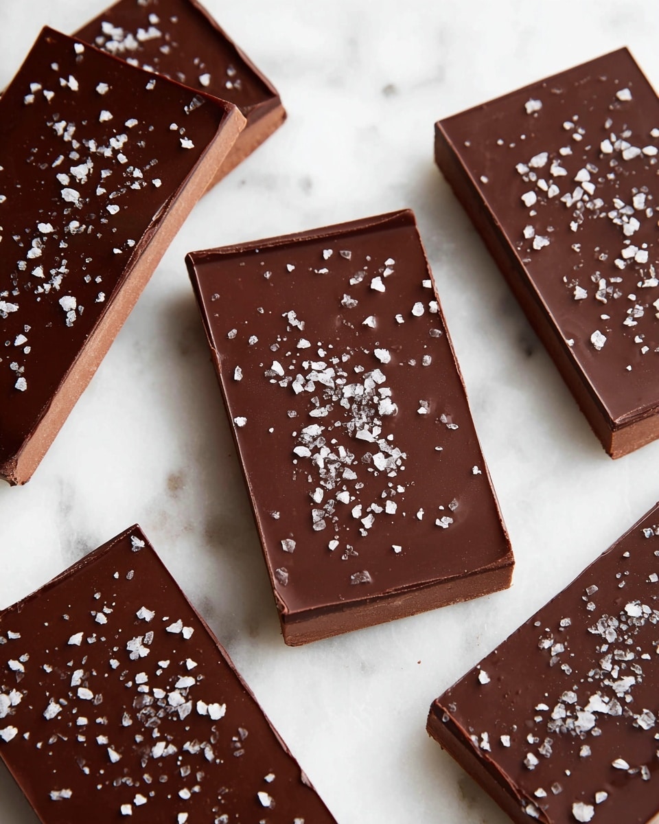 The image shows several rectangular chocolate bars placed on a white marbled texture. Each bar has two visible layers: a thicker bottom layer in a light brown shade, and a smooth, shiny dark brown top layer. The top layer is decorated with small flakes of white sea salt scattered evenly across the surface. The bars have clean, sharp edges and the texture of the bottom layer looks solid but slightly soft. The photo taken with an iphone --ar 4:5 --v 7