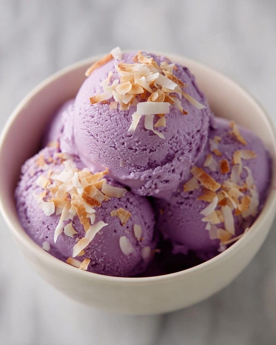 The image shows three scoops of purple ice cream layered closely together in a white bowl. The ice cream has a smooth yet slightly textured surface. On top of the scoops, there is a sprinkle of toasted coconut flakes in light brown and white colors, adding a crunchy texture contrast. The bowl sits on a white marbled surface which gives a clean and bright background. photo taken with an iphone --ar 4:5 --v 7