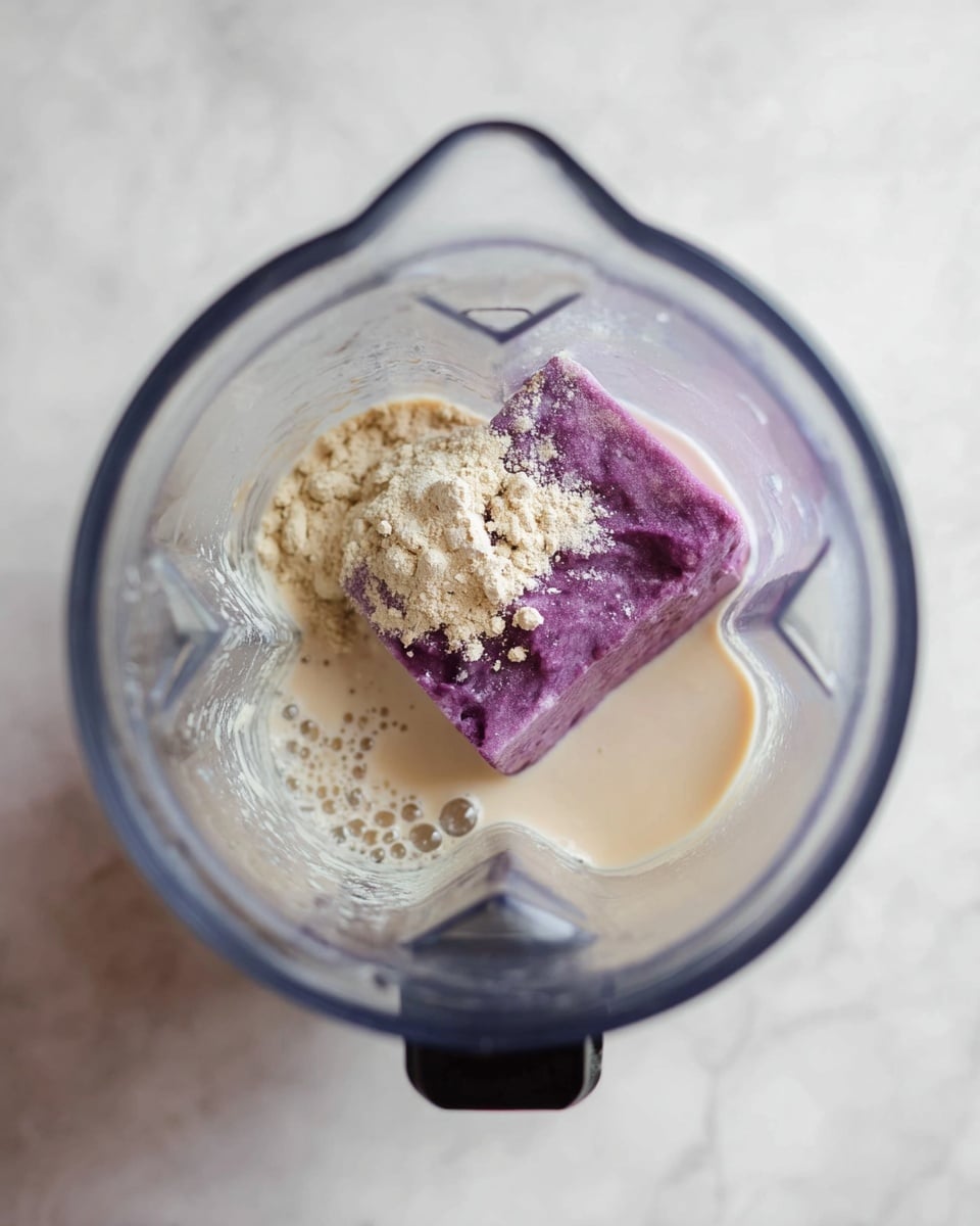 Inside a clear blender container on a white marbled surface, there are three main layers: at the bottom, a small pool of light beige liquid, likely a type of milk or cream, with some bubbles around the edges; in the middle, a thick, bright purple block that looks like frozen fruit or a smoothie base; and on top of the purple block, a light tan powder unevenly spread, some in small clumps. The blender’s top is black and partially visible, and the scene is clean and simple. photo taken with an iphone --ar 4:5 --v 7