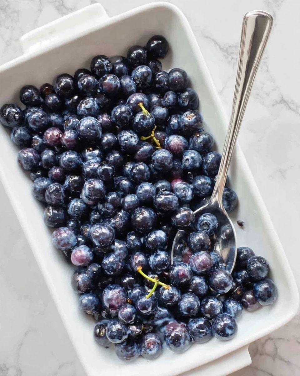 A close-up view of a white rectangular dish filled with a single layer of fresh dark blue and purple blueberries, with some blueberries showing light natural stems. A silver spoon rests among the blueberries on the right side of the dish. The dish is placed on a white marbled surface. photo taken with an iphone --ar 4:5 --v 7