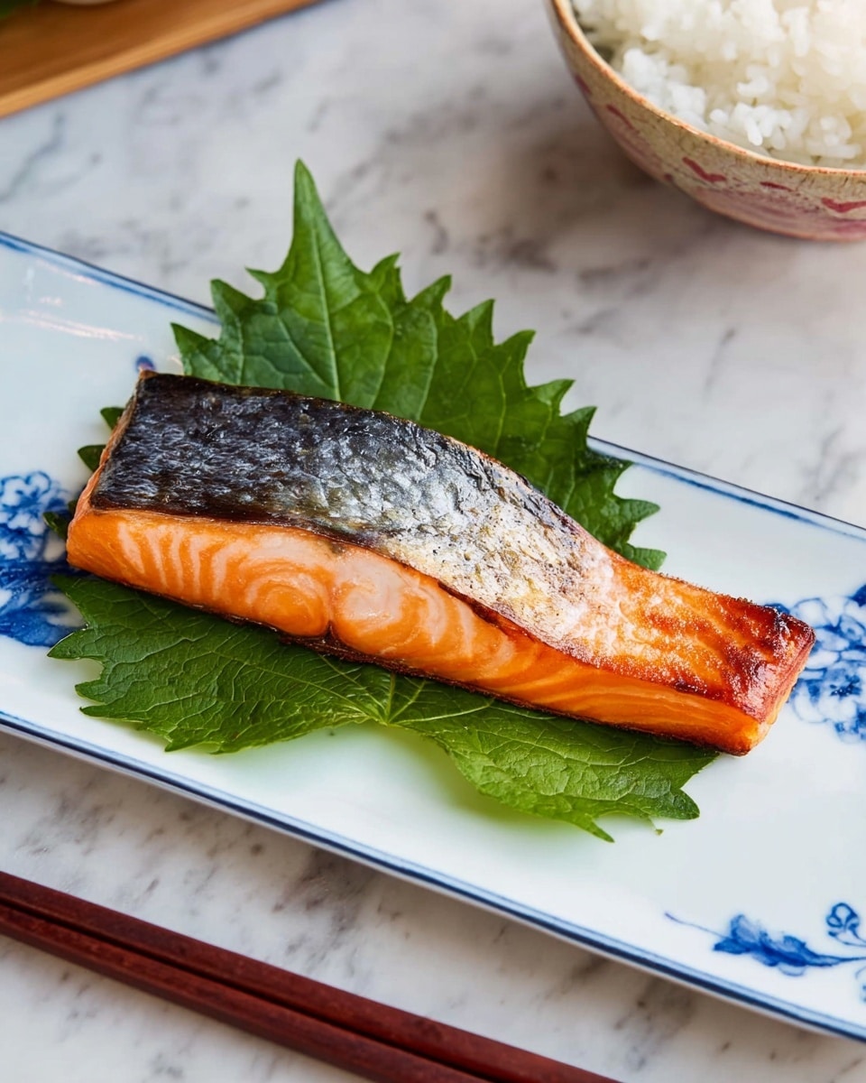 A piece of grilled salmon with crispy dark grey skin on top and bright orange tender flesh below is placed on two large green shiso leaves. The fish rests on a white rectangular plate with blue decoration on one end, which sits on a white marbled surface. Part of a bowl with white rice is visible in the top right corner, and wooden chopsticks rest next to the plate, held by a woman's hand. Photo taken with an iphone --ar 4:5 --v 7