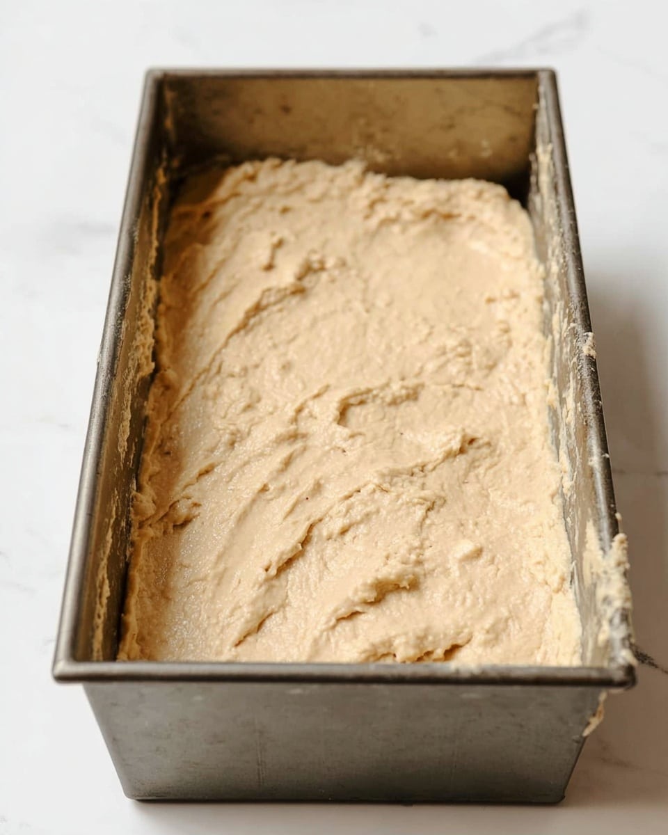 The image shows a metal rectangular baking pan filled with an uneven layer of light beige, thick batter spread inside it. The batter has a slightly rough texture with small peaks and ridges visible across the surface, indicating it has been spooned or poured in but not yet baked. The pan itself looks a bit worn with some batter stuck near the edges. The pan is placed on a white marbled surface, with a clean and bright background. Photo taken with an iphone --ar 4:5 --v 7