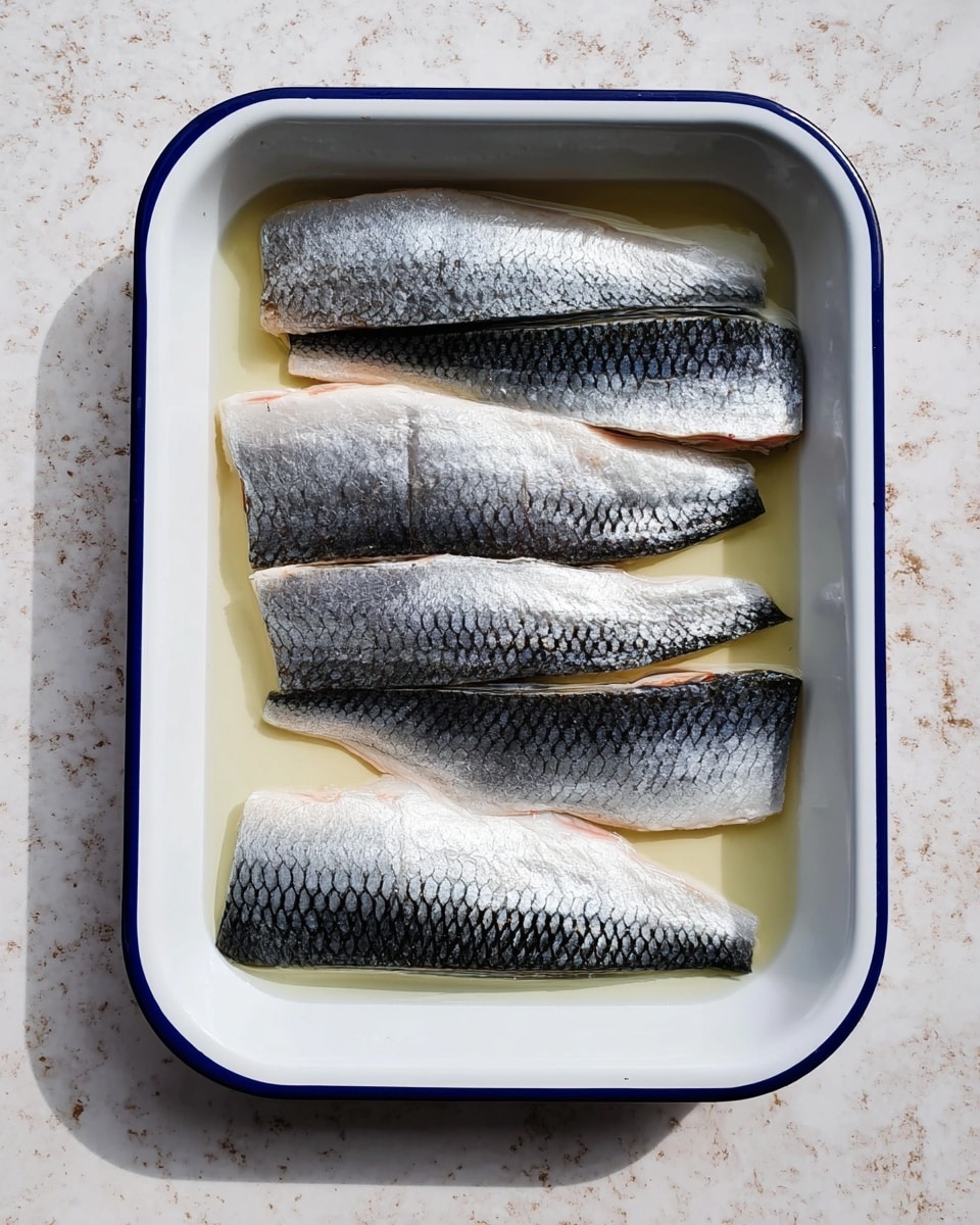 A white rectangular tray with a blue rim holds several pieces of raw fish fillets neatly placed side by side. Each fillet has a shiny, silver skin with a dark, scaled pattern in the middle, and a lighter, smooth underside. The fillets vary slightly in size but all lie flat in the tray, partially submerged in a clear liquid. The tray is set on a white marbled surface with soft natural light casting gentle shadows. photo taken with an iphone --ar 4:5 --v 7