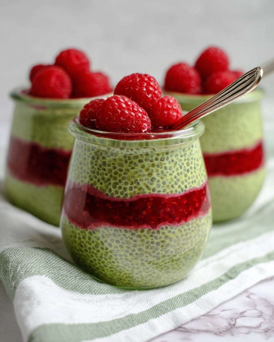 The image shows three small glass jars filled with two layers of chia pudding, placed on a white marbled surface with a white cloth that has green stripes. The bottom layer in each jar is bright red and has a jelly-like texture with visible raspberry pieces. The top layer is light green with a thick, slightly bumpy texture full of chia seeds. Each jar is topped with three whole fresh raspberries that add a bright red contrast. One jar in the front has a spoon inside it. The background is soft and out of focus, highlighting the jars in the front. photo taken with an iphone --ar 4:5 --v 7