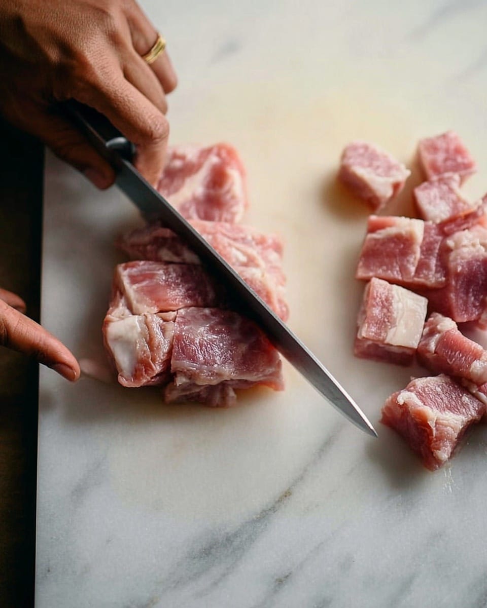 The image shows a close-up of raw meat being sliced on a white cutting board with a white marbled texture beneath. There are two clusters of the same raw meat: one block is being cut into thick rectangular pieces while the other cluster is already cut into smaller cubes. A woman's hand is steadying the block of meat as a knife slices through it. The meat is pinkish with patches of white fat visible inside. The scene is simple and focused on the precise cutting process. photo taken with an iphone --ar 4:5 --v 7