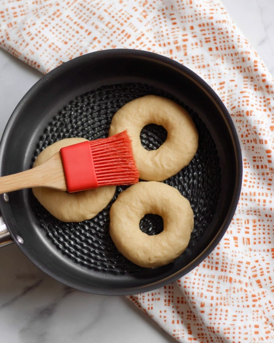 The image shows three uncooked circular dough pieces with holes in the center placed inside a black round perforated pan. A red brush with a wooden handle is applying a coating on the dough. The pan rests on a white marbled surface with a white cloth with orange square patterns underneath. Photo taken with an iphone --ar 4:5 --v 7