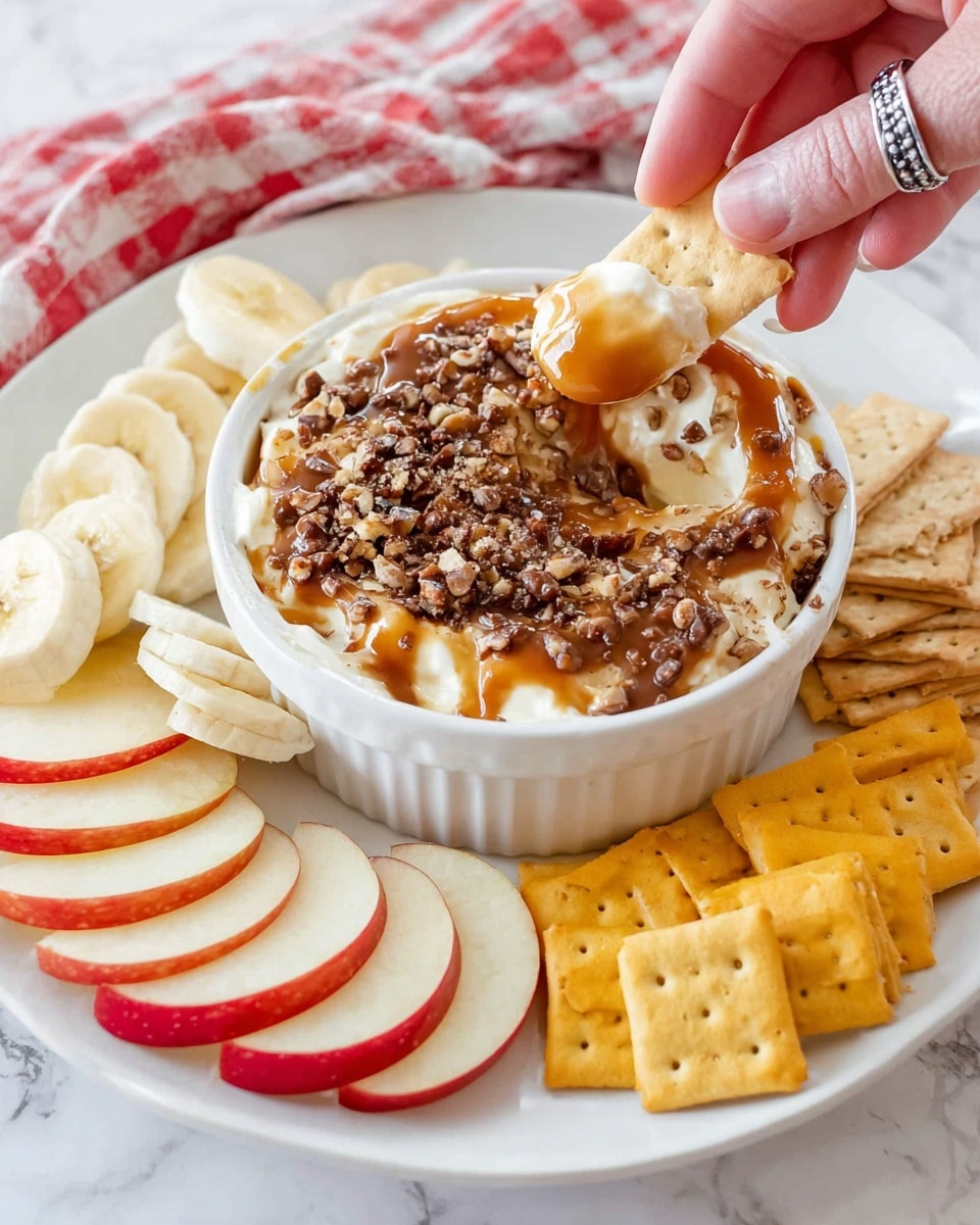 The image shows a white plate with a small white bowl at the top, filled with layers of soft cream cheese, a thick light brown caramel drizzle, and sprinkled dark brown toffee bits on top. A woman's hand with a silver ring is dipping a light tan cracker into the bowl, scooping up the creamy mix. Around the bowl on the plate are several slices of red apple with white flesh, a pile of round banana slices with pale yellow color, yellow square crackers, and light brown rectangular crackers. The plate sits on a white marbled surface with a red and white checkered cloth partially visible in the background photo taken with an iphone --ar 4:5 --v 7