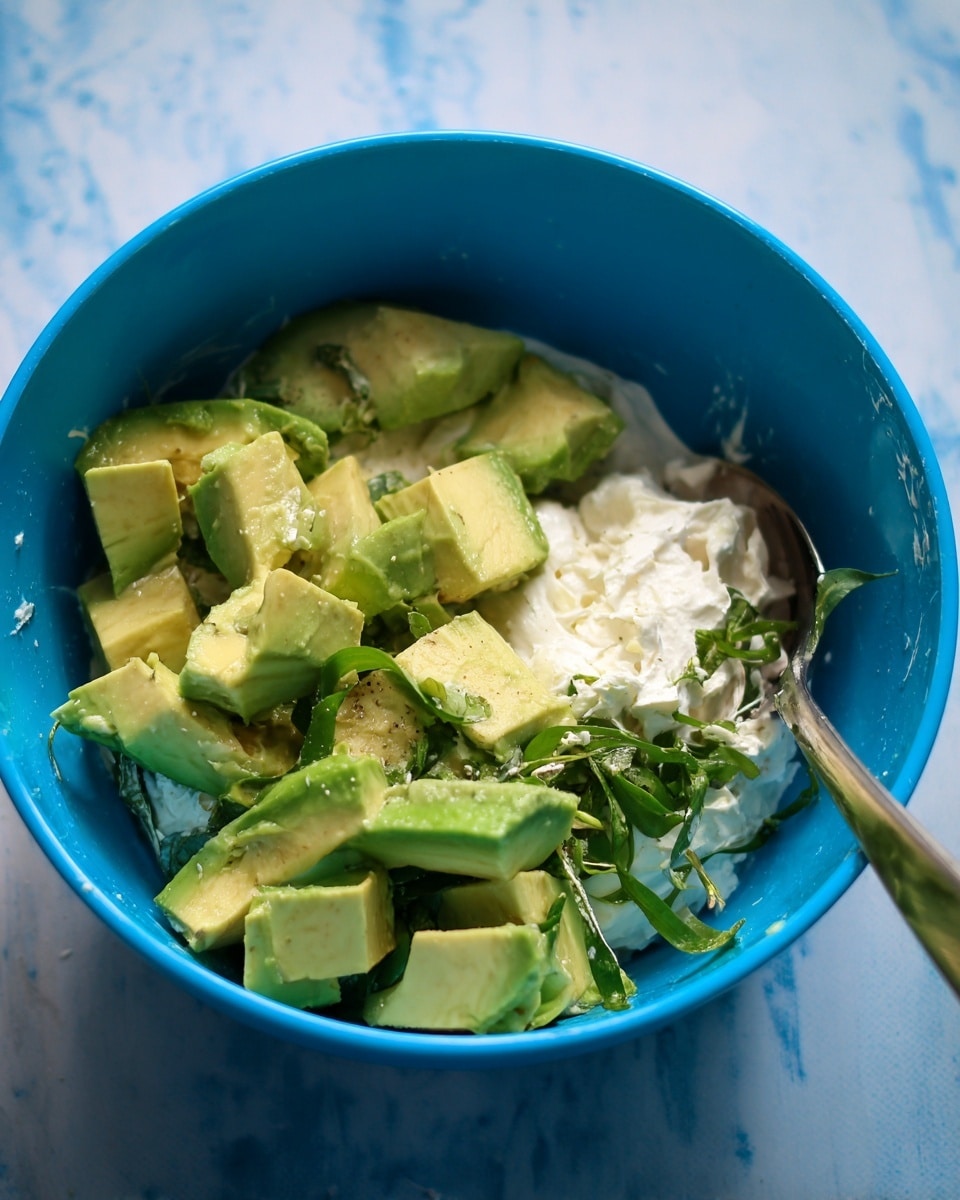 The image shows a bright blue bowl filled with layers of food. At the bottom, there is a white creamy layer, topped with big pieces of fresh green avocado, sliced in a grid pattern but staying in chunks. There is also some green leafy herb mixed in, adding a fresh touch. A silver spoon is resting inside the bowl on the right side. The bowl is placed on a white marbled surface. photo taken with an iphone --ar 4:5 --v 7