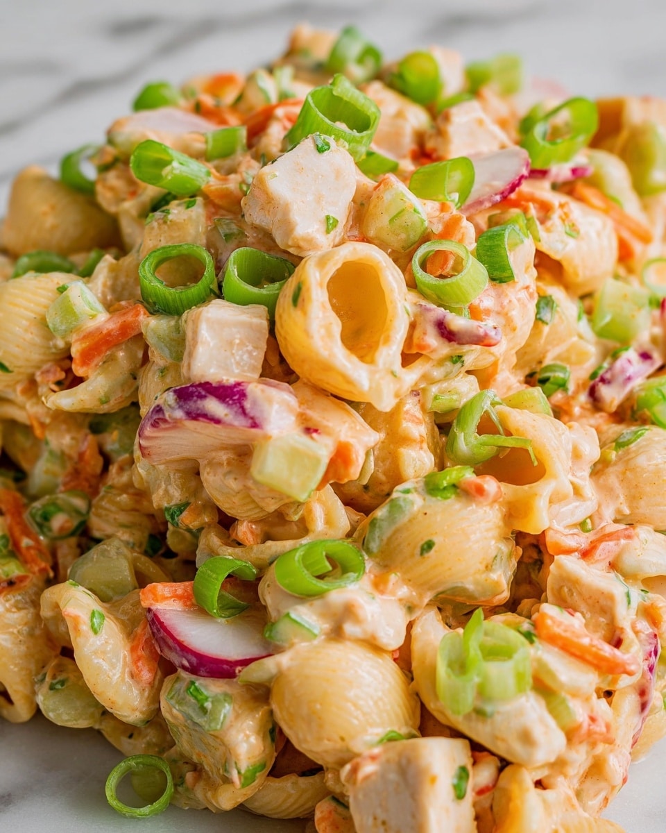 The image shows a close-up of a creamy pasta salad with about four layers visible in texture and color. The main layer consists of short shell pasta coated in a light orange, smooth sauce. Mixed with the pasta are chunks of white chicken pieces, small diced orange carrots, light green celery, and thin purple-red slices of radish, adding texture and color contrast. Scattered on top are bright green rings of chopped green onions that add freshness and a crisp look. The entire dish is placed on a white marbled surface. Photo taken with an iphone --ar 4:5 --v 7