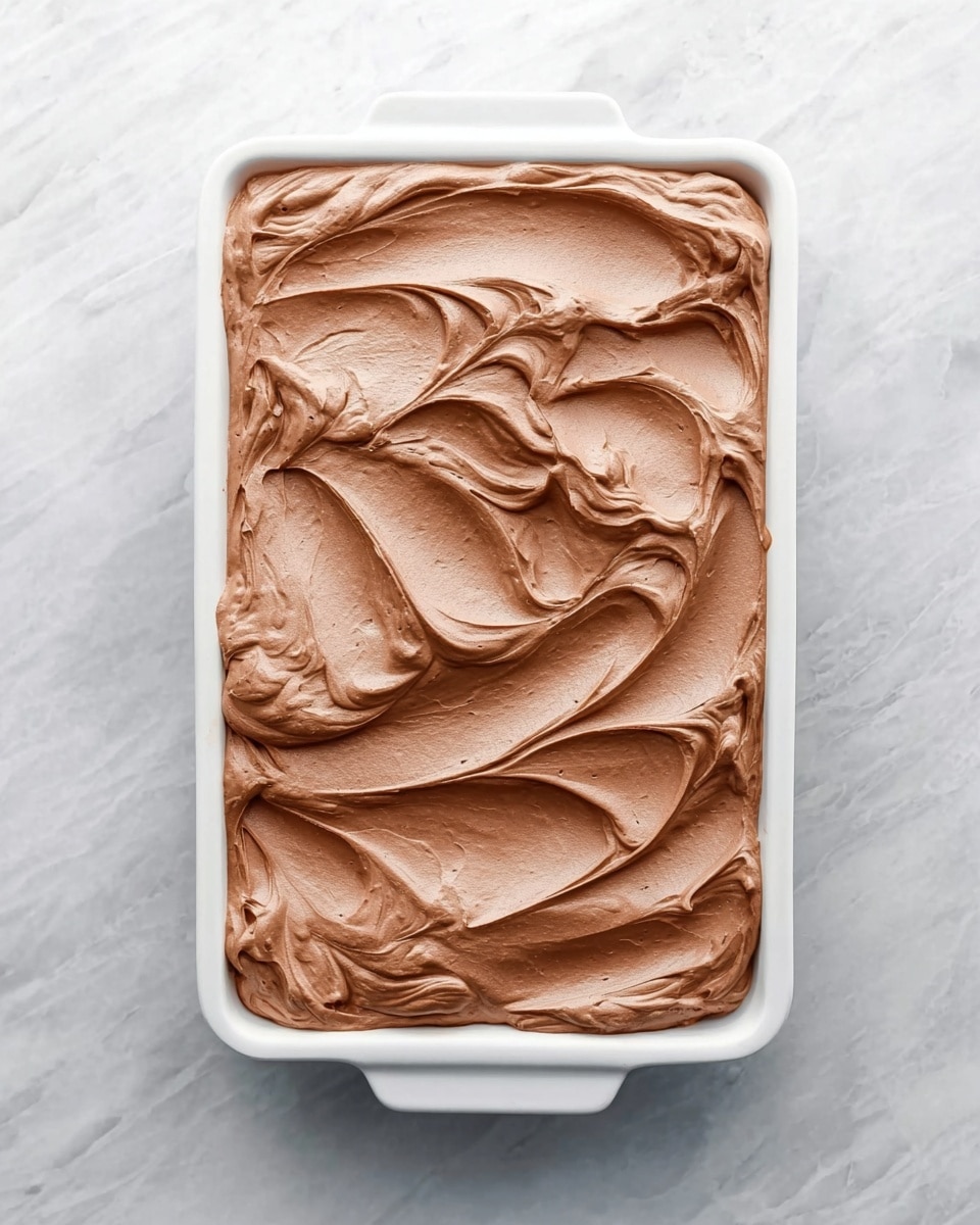 The image shows a white rectangular dish filled with a thick layer of smooth, light brown chocolate mousse spread evenly across the surface with swirled, creamy texture and soft peaks. The dish is placed on a white marbled surface, giving a clean and bright look to the photo taken with an iphone --ar 4:5 --v 7