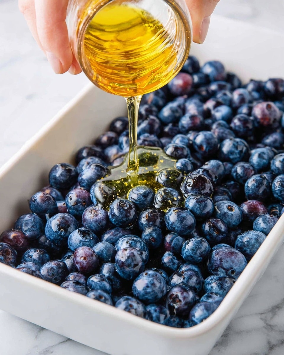 A close-up view of a white rectangular dish filled with fresh blueberries that are plump and deep blue with some having a light shine. Above the blueberries, a woman's hand is tilting a small clear glass container, pouring golden honey that flows softly over the berries, creating a sticky, shiny layer on top. The background is a white marbled surface. photo taken with an iphone --ar 4:5 --v 7